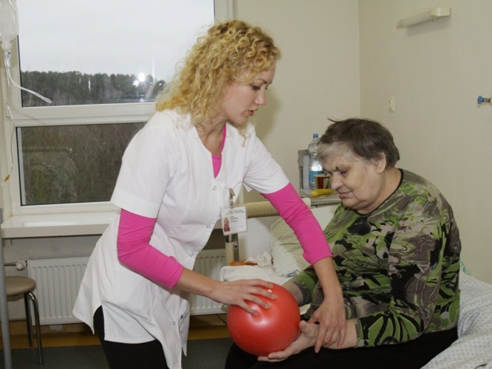 Geriatrijos skyriuje kineziterapeutė Ramunė Matiušovienė padeda pacientei atlikti pratimą su masažiniu kamuoliu. Medikė kasdien ligoniams pritaiko specialius pratimus pagal žmonių judėjimo galimybę, kad pagerintų jų kasdienę veiklą. 